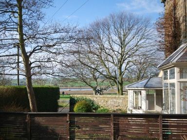 A garden with trees and bushes next to a glass conservatory with a river and fields in the background at Beach House (Alnmouth) in Alnmouth