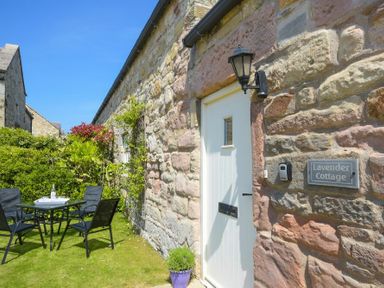 A stone exterior wall with a white door and a black outdoor light next to a garden table and chairs at Lavender Cottage Village Farm in Seahouses