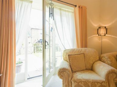A beige armchair with a cushion next to glass doors with white and orange curtains and a floor lamp at Lavender Cottage Village Farm in Seahouses