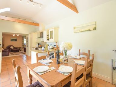 A dining area with a wooden table set with plates glasses and a vase of flowers next to a kitchen with cream cabinets and terracotta tile floor at Lavender Cottage Village Farm in Seahouses