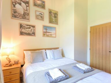 A bedroom with a double bed with pillows and towels on top a wooden bedside table with a lamp six framed pictures on the wall and a wooden door at Lavender Cottage Village Farm in Seahouses