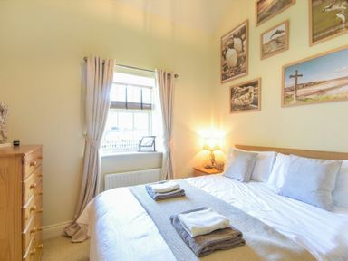 A bedroom with a bed with pillows and folded towels window with curtains a wooden chest of drawers and framed pictures on the wall at Lavender Cottage Village Farm in Seahouses