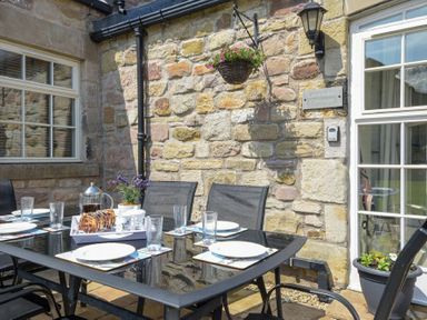 An outdoor dining area with a glass table set with plates and glasses against a stone wall at Lavender Cottage Village Farm Seahouses