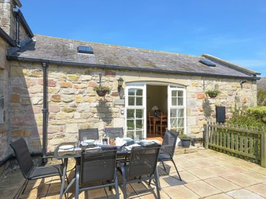 An outdoor patio with a dining table and chairs in front of a stone building with open double doors at Lavender Cottage Village Farm in Seahouses