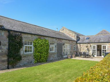 An outdoor patio area with a lawn and stone buildings with windows and climbing plants at Lavender Cottage Village Farm in Seahouses
