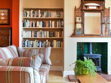 A living room with a striped sofa a bookshelf filled with books a wood stove with green tiled surround and a wooden mirror above at Estuary View in Alnmouth