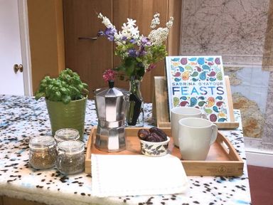 A kitchen table with a green potted plant three spice jars a silver coffee maker a vase with flowers two white mugs a bowl with dates and a cookbook with a colorful cover at Estuary View in Alnmouth