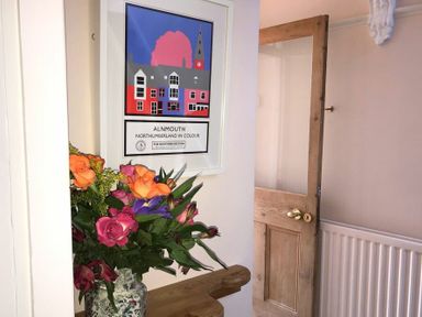 A hallway with a wooden door radiator a framed Alnmouth poster and a vase of flowers on a ledge at Estuary View in Alnmouth