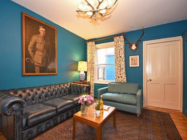 A living room with a leather sofa a small couch a wooden coffee table a lamp a window with curtains a painting and antlers on the wall at Estuary View in Alnmouth