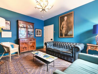 A living room with a blue leather sofa a wooden cabinet a wicker chair and a framed portrait at Estuary View in Alnmouth