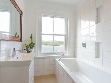 A bathroom with a white bathtub a sink with plants on the counter and a window showing a landscape at Estuary View in Alnmouth