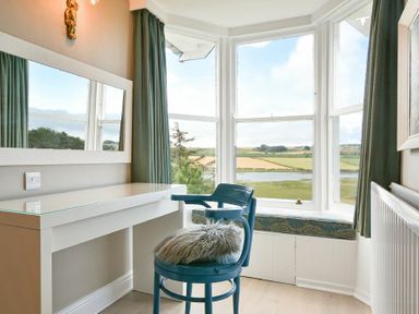 A room with a blue chair and a white desk next to bay windows with green curtains and a window seat at Estuary View in Alnmouth