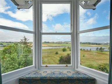 A bay window with a cushioned seat overlooking fields and a river at Estuary View in Alnmouth