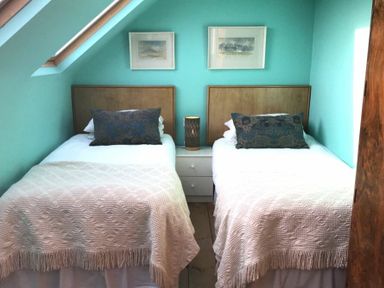 A bedroom with two single beds white blankets and pillows a nightstand with a lamp and pictures on the turquoise wall at Estuary View in Alnmouth