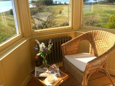 A wicker chair with a cushion next to a small table with a cup, glasses, a newspaper, and a vase of flowers by windows overlooking a grassy area at Estuary View in Alnmouth