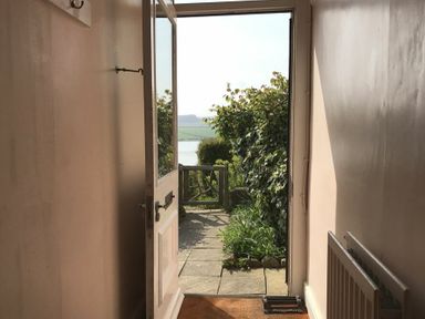 A hallway with an open door leading to a garden with plants and a view of fields and water at Estuary View in Alnmouth