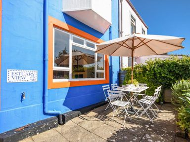 An outdoor patio with a white table and chairs under a large umbrella next to a blue building with an orange window frame at Estuary View in Alnmouth