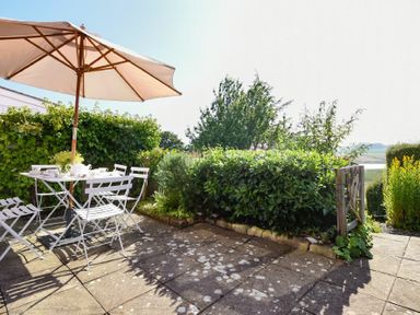 A patio with a white table and chairs under a large beige umbrella surrounded by green bushes at Estuary View in Alnmouth