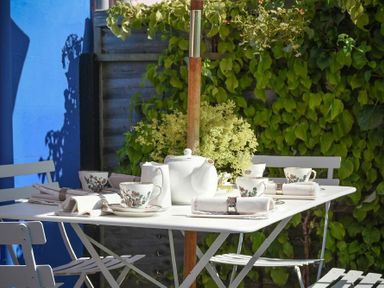 An outdoor white table set with a teapot cups napkins and cutlery surrounded by white chairs and green foliage in the background