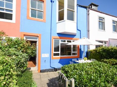 A blue and orange house exterior with white windows and an outdoor patio set with umbrella surrounded by bushes at Estuary View in Alnmouth