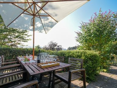 An outdoor patio with a wooden table and chairs under a large umbrella with glassware and a bottle on the table at Estuary View in Alnmouth