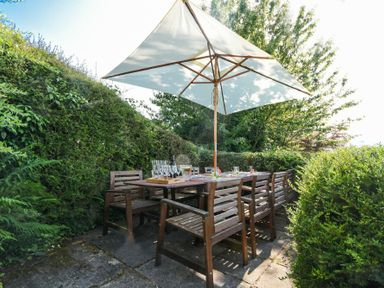 An outdoor patio dining area with wooden chairs and a table under a large umbrella surrounded by greenery at Estuary View in Alnmouth