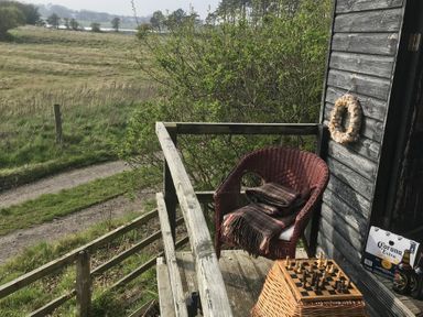 A balcony with a wicker chair and blanket a wicker table with a chess board and pieces a wreath hanging on a wooden wall and a box of Corona Extra beer at Estuary View in Alnmouth