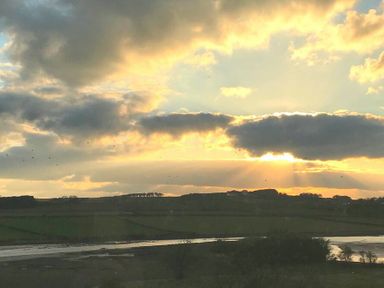 A landscape showing a river with fields and hills in the background under a cloudy sky with sun rays at Estuary View in Alnmouth