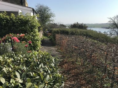 A garden path with bushes and flowers next to a white building and a river with boats in the background at Estuary View in Alnmouth