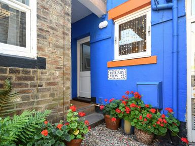 The entrance of a blue-painted building with orange trim and flower pots with red flowers at Estuary View in Alnmouth