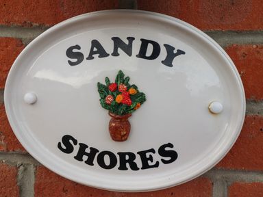 An oval white plaque with the words sandy shores and a flower vase mounted on a brick wall