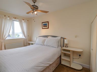 A bedroom with a bed a ceiling fan two bedside tables with lamps a window and curtains at Sandy Shores in Seahouses