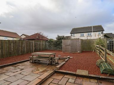 A backyard with wooden picnic table and bench on stone patio next to shed and fenced garden at Sandy Shores in Seahouses