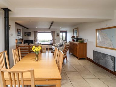 A dining table with chairs and a vase of yellow flowers next to a sitting area with sofas and a wooden sideboard with a plant and decorations at Kittling Cottage in Bamburgh