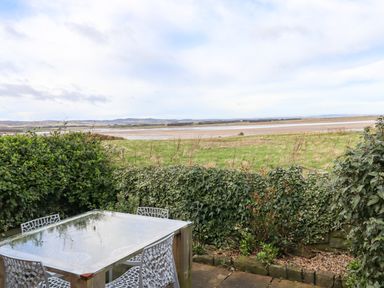An outdoor patio with a glass table and four chairs next to a hedge with a grassy field and water in the background at Kittling Cottage in Bamburgh