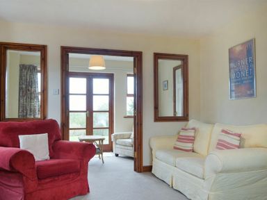 A living room with a red armchair a white sofa with striped pillows wooden framed mirrors and a view into a room with glass doors at Smithy Court in Craster
