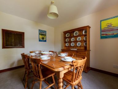 A dining room with a wooden table set with white dishes and glasses with wooden chairs and a wooden cabinet displaying decorative plates at Smithy Court in Craster