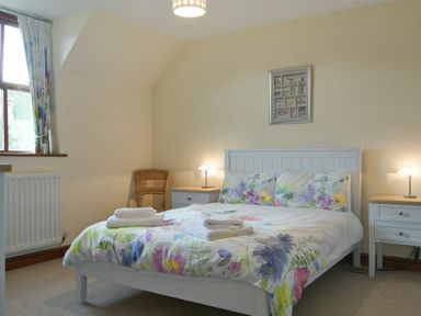 A bedroom with a double bed floral bedding two bedside tables with lamps a wicker chair and a window at Smithy Court in Craster