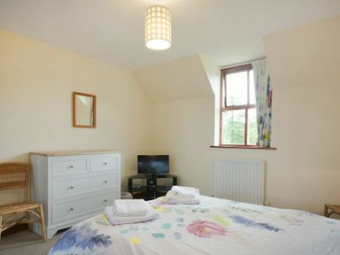 A bedroom with a bed with floral bedding towels on the bed a white dresser a small TV on a stand a window with patterned curtains and a wicker chair at Smithy Court in Craster