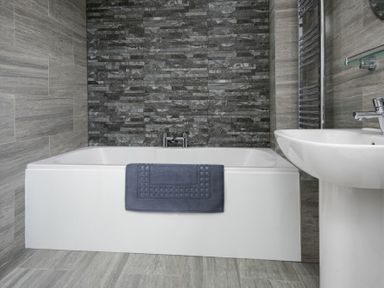 A bathroom with a white bathtub a blue towel hanging over it a pedestal sink and gray tiled walls at Smithy Court in Craster
