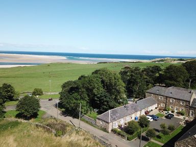 Stone buildings with parked cars near a grassy field and beach at Lapwing Cottage in Bamburgh