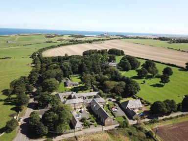 An aerial view of a rural area with several stone buildings surrounded by trees fields and a distant view of the sea at Lapwing Cottage in Bamburgh