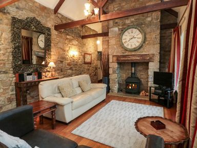 A living room with a stone fireplace a clock above and a cream sofa at Lapwing Cottage in Bamburgh