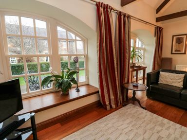 A living room with arched windows with plants on the windowsill a black leather sofa a wooden side table and striped curtains at Lapwing Cottage in Bamburgh