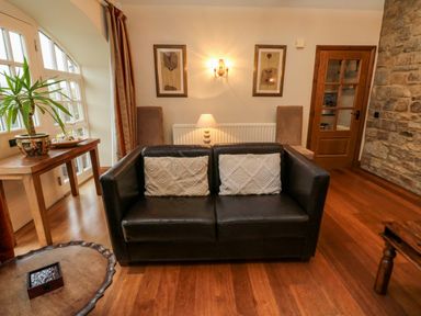 A living room with a black leather sofa with cushions a wooden table with a plant framed pictures on the wall and wooden floor at Lapwing Cottage in Bamburgh