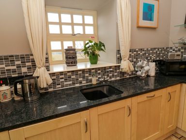 A kitchen with a black countertop a sink a window with curtains and a mosaic tile backsplash at Lapwing Cottage in Bamburgh