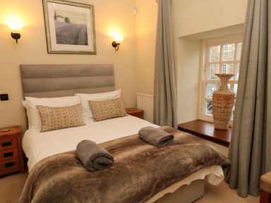 A bedroom with a double bed two rolled towels bedside tables a window with curtains and a large wicker vase at Lapwing Cottage in Bamburgh