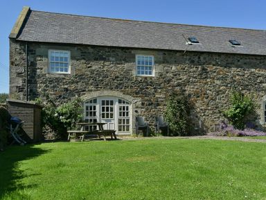 An exterior view of a stone house with grass lawn and outdoor furniture at Budle Sands in Bamburgh