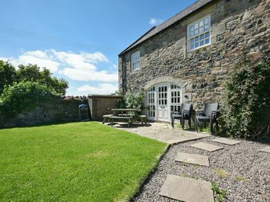 A stone house with white windows and doors a green lawn a wooden picnic table black chairs and a gravel path at Budle Sands in Bamburgh