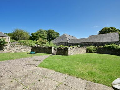 A stone paved patio leading to a grass lawn with a stone wall and traditional stone buildings in the background at Budle Sands in Bamburgh
